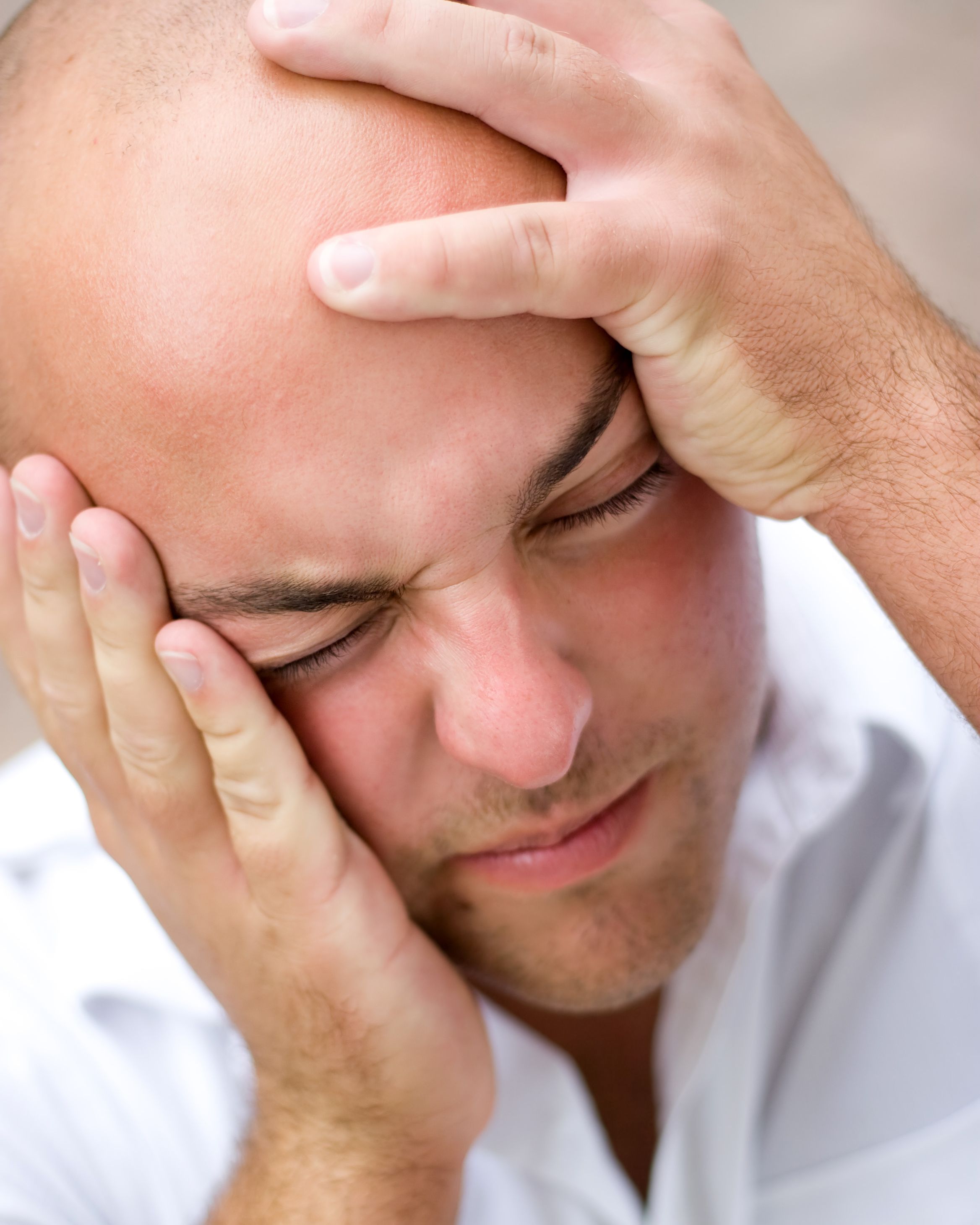 A man in a white shirt holds his head with both hands, eyes closed in apparent discomfort, suggesting a headache or stress-related pain. The clean, neutral background and professional lighting make this suitable for educational posts about headache management, stress relief techniques, or general pain management topics that would resonate with the practice's focus on comprehensive family care.