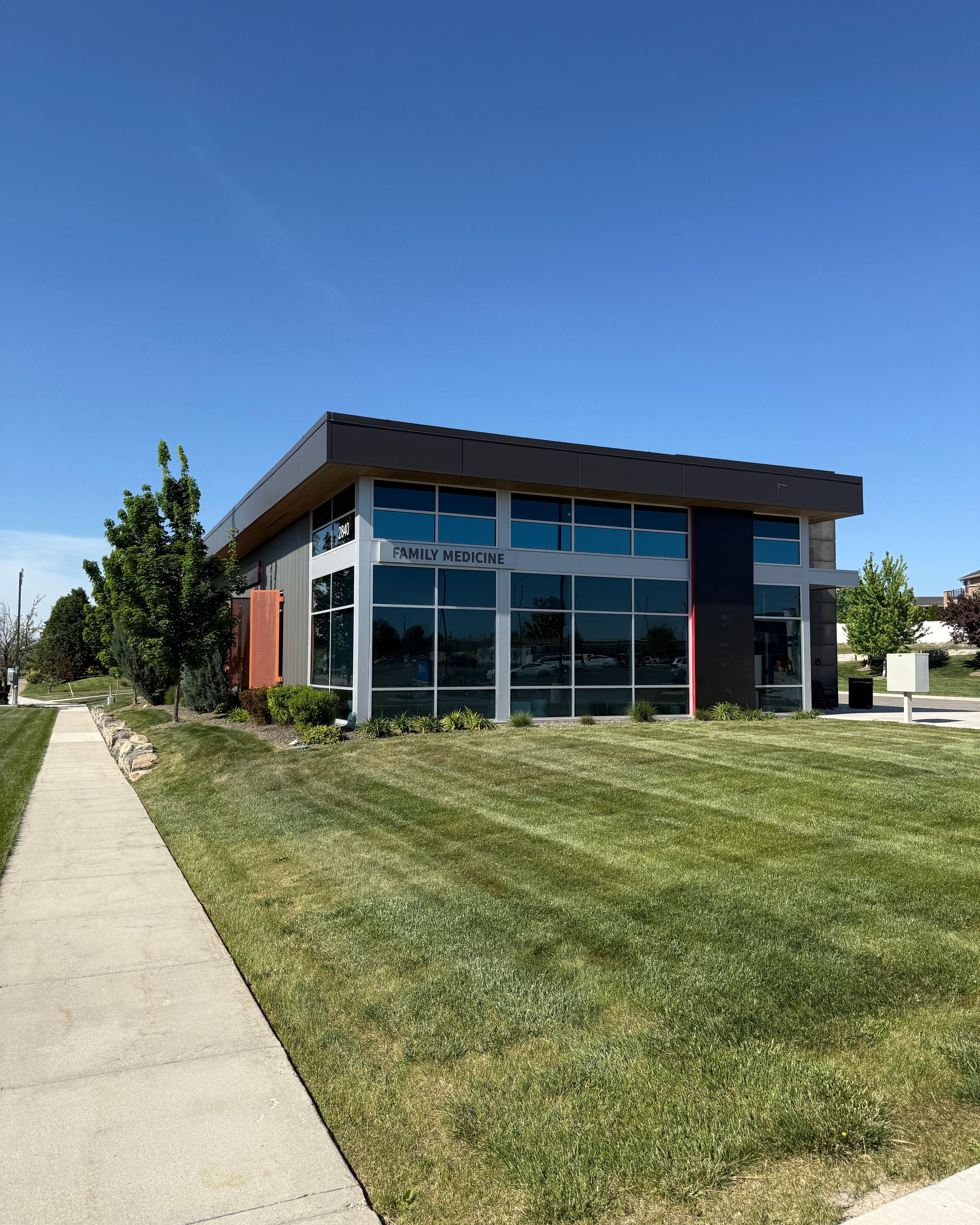 This image shows the modern, contemporary exterior of the Ridgeview Family Health clinic building on a clear, sunny day. The two-story medical facility features large glass windows, clean architectural lines with dark and light contrasting materials, and well-maintained landscaping with green lawn and trees surrounding the property. This professional, welcoming exterior shot would work well for posts about the practice's location, accessibility, or general practice information, conveying a sense of modern, quality healthcare in a comfortable community setting.