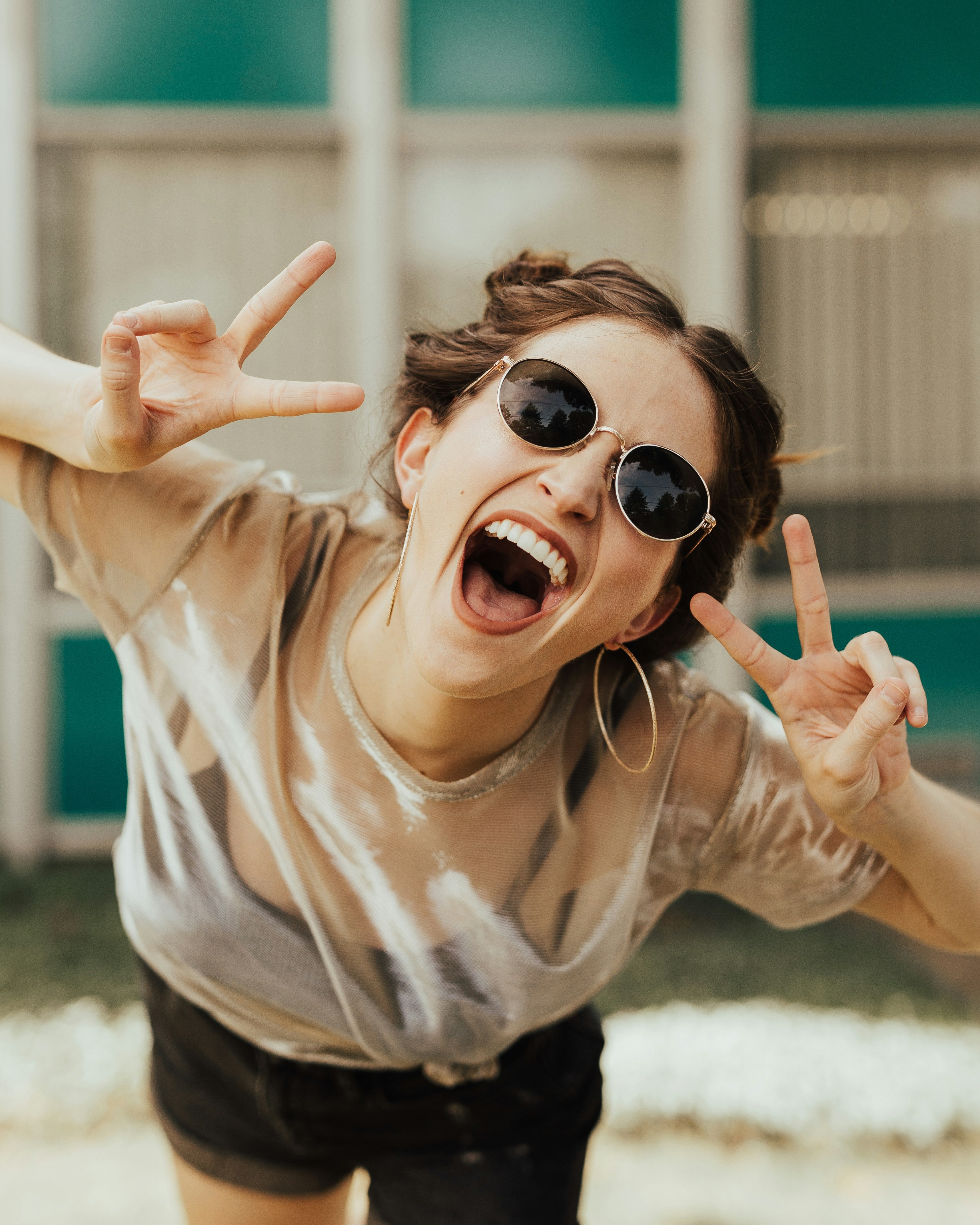A young woman with brown hair in a messy bun wears round sunglasses, gold hoop earrings, a sheer beige top, and black shorts while striking a playful pose with both hands making peace signs and her mouth open in an excited laugh. She's photographed outdoors against a blurred green and white building facade, giving the image a bright, energetic summer vibe. This upbeat, high-quality lifestyle shot would work well for social posts about young adult or women's health, accepting new patients, or lighthearted seasonal community content.